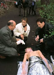 Die Leiche eines Jungen (Komparse, lieg.) ist gefunden worden. Der Gerichtsmediziner (Klaus Schindler, li.) spricht mit der Staatsanw&auml;ltin (Britta Schmeling) und Ralf Bongartz (Max Gertsch) &uuml;ber eine m&ouml;gliche Todesursache.