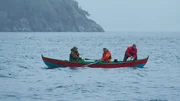Drei M&auml;nner in einem Boot fahren durch neblige Gew&auml;sser nahe steiler K&uuml;sten.