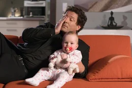 Christoph Baumgartner (Francis Fulton-Smith) sieht mit dem Baby Rosi eine Sendung im TV, in der seine Freundin Chantal interviewt wird.