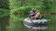 Zwei Personen sitzen in einem Schlauchboot auf einem ruhigen Fluss, umgeben von &uuml;ppiger gr&uuml;ner Vegetation.