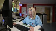 In Magdeburg a middle aged blonde nurse is working at a desk in the staff room. She is wearing glasses and is looking intently at the screen, while her right hand lays on the mouse. Behind her we see a red wall, a big radiatior, a big metallic box among other things.
