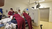 Berlin: four members of staff, three nurses wearing dark red scrubs and a radiology assistant wearing a white uniform are moving a patient from the gurney to the CT device. The male nurse is wearing a mask, and all of them are looking down at the patient.