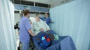 Saarbr&uuml;cken: Physician Sabine in blue scrubs is standing next to patient in hospital bed, talking to him. Patient has a hospital gown on and his clothes are lying on the bed. On his right side nurse Sabrina is handling his IV. On both side are blue curtains.