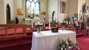 Innenraum einer Kirche mit Altar und einem wei&szlig; bedeckten Tisch, geschm&uuml;ckt mit Blumen und einer Urne w&auml;hrend eines Gottesdienstes.