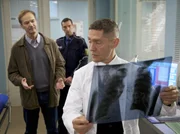 Dr. Rolf Kaminski (Udo Schenk, rechts) traut seinen Ohren nicht, als Oberkommissar Erich Gassmann (Arnfried Lerche, links) ihm sagt, dass ein Patient Anzeige gegen ihn erstattet hat. Kaminski hat Sylvie Meinert davor bewahrt von Walter Hahn geschlagen zu werden, es ist zu einem Handgemenge zwischen den beiden M&auml;nnern gekommen und dabei ist es bei Walter zu einer Nachblutung seiner frischen OP-Wunde gekommen. Walter Hahn hat bei den beiden Polizisten (Komparse, Mitte) angegeben, dass Kaminski ihn angegriffen h&auml;tte. Als der Arzt dem Oberkommissar die Situation aus seiner Sicht schildert, wird deutlich, dass Gassmann nichts von den "G&ouml;ttern in wei&szlig;" h&auml;lt - ein Wort ergibt das andere, bis der Oberkommissar Kaminski verhaftet.