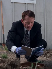 Detective squatting by dug out hole, reading a notebook that he found.
