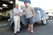 Wide shot of 2CV in front of workshop with Mike and seller standing in front of it and shaking hands