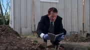 Detective squatting by dug out hole, reading a notebook that he found.