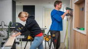 Sherry Holmes (left) uses a chop saw to cut dowel board to length as Michael Holmes (right) tacks it to the wall with a brad nailer as seen on HGTV&rsquo;s Holmes Family Rescue Season 2 Episode 14.