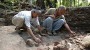 Zwei Personen graben sorgf&auml;ltig aus und untersuchen Keramikfragmente an einer arch&auml;ologischen Ausgrabungsst&auml;tte in einem bewaldeten Gebiet.