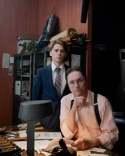 Zwei Personen stehen an einem Arbeitstisch, der mit Werkzeugen, Papieren und Skizzen bedeckt ist, in einer Werkstatt mit Regalen voller Materialien im Hintergrund.