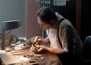 Eine Person sitzt an einer Werkbank voller Werkzeuge und Lederst&uuml;cke und arbeitet sorgf&auml;ltig an einem Schuh.