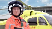 Eine Frau mit Fliegerhelm und orange-schwarzer Uniform vor einem Hubschrauber