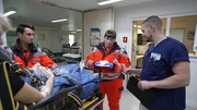 L&uuml;beck: a pixelated female patient has just been brought to the A&E. Nurse Jacob is smilling and seems to be making the handover with two young paramedics, who are both wearing orange uniforms. They are at the A&E entrance. Behind them we see the staff room with a glass window and in the background several empty hospital gurneys. The female paramedic seems to be reading a note in her gloved hands.