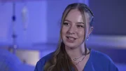 Kaufbeuren: close shot of young nurse Laura-Sophie, who is smiling while giving an interview on the interview set. The background is illuminated in shades of blue. She is wearing earrings and two necklaces.