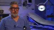 Saarbr&uuml;cken: close shot of a bearded male nurse with sight glasses during an interview in the show's interview set. The light in the background is blue, a big lamp is lit in the background to the right and hovering over a hospital bed covered with a white sheet. He is wearing blue scrubs and has a telephone in his pocket.