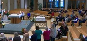 In der Kirche steht w&auml;hrend des Trauergottesdienstes ein mit Blumen umgebener Sarg, w&auml;hrend die Anwesenden in den B&auml;nken sitzen oder stehen und aus ihren Heften lesen.