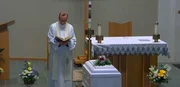 Ein Priester steht vor dem Altar in der Kirche und h&auml;lt ein ge&ouml;ffnetes Buch, w&auml;hrend vor ihm ein von Blumen umgebener Sarg steht.