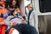 Mit dem Schrecken davongekommen: Im Mordfall an einem Textilproduzenten, in den auch die Schwester von Kriminalkommissarin Lea Gomez verwickelt ist, wird pl&ouml;tzlich einer ihrer Mitarbeiter angeschossen. Wollte die Tatperson erneut zuschlagen? Die Rettungssanit&auml;ter (Komparsen, l.) versorgen Robin F&uuml;hrich (Steven Preisner, M.), w&auml;hrend Kriminalhauptkommissarin Martina Seiffert (Astrid M. F&uuml;nderich) bereits zu eruieren versucht, in welchem Zusammenhang die beiden Vorf&auml;lle stehen k&ouml;nnten.