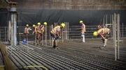 Auf der Baustelle im Inneren des Hochhauses verlegen Bauarbeiter millimetergenau tonnenschwere Stahlarmierungen.