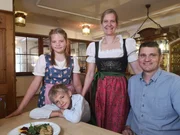 Familie Bauer von "Der Obere Wirt zum Queri" in Frieding-Andechs sitzen und stehen an einem Tisch in der Gaststube. Auf dem Tisch steht ein Tellergericht.
