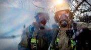 Zwei Einsatzkr&auml;fte der Feuerwehr Duisburg mit Atemschutzmasken auf einem verrauchten Gel&auml;nde. Eine schwarze Rauchwolke steigt &uuml;ber einem verlassenen Vereinsgel&auml;nde auf, als der L&ouml;schzug der Wache 3 eintrifft.