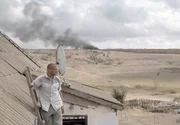 Tolik (Sergiy Shadrin) steht auf einer an sein Hausdach gelehnten Leiter und blickt auf die Auswirkungen der K&auml;mpfe in der Umgebung. Im Hintergrund ist die riesige Rauchfahne nach dem Abschuss der malaysischen Verkehrsmaschine MH17 zu sehen, der international f&uuml;r Entsetzen sorgt.