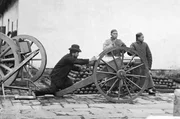 John Thomson (li.) posiert an einer Gatling-Kanone, bereit die Waffe abzufeuern. Neben ihm stehen zwei chinesische M&auml;nner, von denen einer den Verschluss der Waffe h&auml;lt.