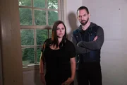 Tight shot of Nick, with arms crossed, and Katrina in front of interior window.