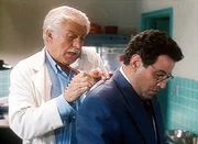 Dr. Mark Sloan (Dick Van Dyke, l.) notiert eine wichtige Adresse. Dabei benutzt er Normans (Michael Tucci, r.) R&uuml;cken als Unterlage.