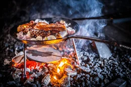 Ein Steak wir &uuml;ber einer offenen Feuerstelle gegrillt wobei eine geschmiedete Eisenpfanne als Schutz vor der direkten Hitze hilf.