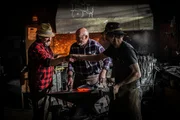 Grillweltmeister Leo Gradl (links) beobachtet Schmied Klaus Wieshofer (Mitte) und seinen Gesellen beim Schmieden einer Eisenpfanne.