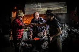 Grillweltmeister Leo Gradl (links) beobachtet Schmied Klaus Wieshofer (Mitte) und seinen Gesellen beim Schmieden einer Eisenpfanne.