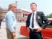 MacGyver (Richard Dean Anderson, l.) und Stringer (Randolph Mantooth, r.) stehen vor dem Hotel, in dem Prometheus einen Brandanschlag ver&uuml;bt hat.