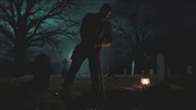 Eine Person steht nachts auf einem Friedhof, von einer Laterne auf dem Boden beleuchtet und h&auml;lt dabei ein Werkzeug &uuml;ber frisch aufgeworfener Erde.
