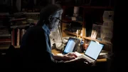 Eine Person sitzt an einem Schreibtisch in einem schwach beleuchteten Raum und wird vom Licht eines Laptops erhellt, w&auml;hrend sie von B&uuml;chern und Unterlagen umgeben arbeitet.