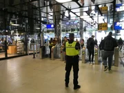 Menno standing in front of the check in ports at Amsterdam Central Station while colleague Baran helps a traveller in the background.