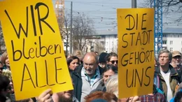 Auf dem gelben Schild in der ausgew&auml;hlten Region steht der deutsche Slogan &bdquo;WIR bleiben ALLE&ldquo;. Dieser Slogan ist ein wichtiger Slogan f&uuml;r Bewegungen f&uuml;r Wohnrechte und gegen Gentrifizierung, insbesondere in Berlin.