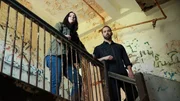 Hosts Katrina Weidman and Nick Groff stand on stairway landing above with decrepit walls behind them.