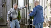 Zwei M&auml;nner stehen vor einem Geb&auml;ude, und einer von ihnen zeigt mit der Hand auf den anderen, w&auml;hrend sie sich neben einem Metallrohr unterhalten.