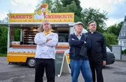 Hans Zielonka (Michael Hanemann, M.), Pastor Puttermann (Johannes Rotter, r.) und Hubert (Matthias Komm, l.) stehen vor Huberts H&auml;hnchenwagen und schauen zum "Pascha Grill", wo die Hindus ihren Gottesdienst feiern.