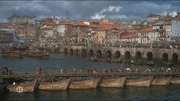 Eine lebendige historische Stadt am Fluss zeigt eine belebte Br&uuml;cke und Boote auf dem Wasser, umgeben von Geb&auml;uden mit roten D&auml;chern unter einem dramatischen Himmel.