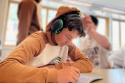 Ein Sch&uuml;ler sitzt mit Kopfh&ouml;rer in der Klasse und versucht sich zu konzentrieren w&auml;hrend der anderen Sch&uuml;ler ihn ablenken.