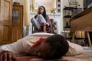 Oliver Cowie (Clarence Smith) liegt mit einer blutenden Wunde unter dem Tisch. Im Hintergrund sitzt seine Frau Lyn Cowie (Mina Andala).