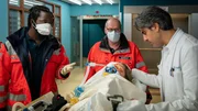 Dr. Schmidt (Joel Williams, l.) &uuml;bergibt Dr. Sharif (Atheer Adel, r.) den verletzten Tommy Riggers (Paul Ahrens, M.), der aus Panik von einem Balkon gesprungen ist.