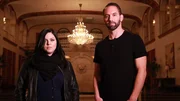 Hosts Katrina Weidman and Nick Groff stand in grand lobby of Statler City Hotel with serious faces.