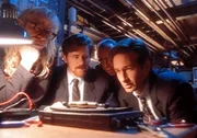Mulder (David Duchovny, r.), Langly (Dean Haglund, l.), Byers (Bruce Harwood, 2.v.l.) und Frohike (Tom Braidwood, 2.v.r.) betrachten einen Kabelfernsehdecoder, der offenbar konstruiert wurde, um das Bewusstsein der Zuschauer zu manipulieren.