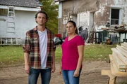 Zwei Personen stehen in einem Hof vor einem Haus und einer Werkstatt mit Werkzeugen und Baumaterialien.