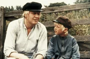 Alfred (Bj&ouml;rn Gustafsson, l.) und Michel (Jan Ohlsson, r.) sitzen auf der Bank und beobachten, wie Lina mit einem fremden Bauern flirtet.
