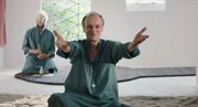 Stella (Edgar Selge) breitet ihre Arme nach vorne aus, w&auml;hrend Roman (Thomas Wodianka) im Hintergrund etwas in einem M&ouml;rser zerreibt.